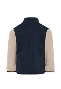 Ζακέτα από φλις με ψηλό γιακά· σκούρο μπλε πλάτη και μανίκια κρεμ, υφασμάτινη υφή και μαύρο τελείωμα στο κάτω μέρος των μανικιών.