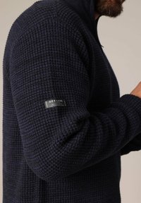 Pull en mailles texturées bleu marine avec un motif côtelé, col zippé mi-long et une étiquette de marque sur la manche gauche. Le tissu a l'air épais et confortable.