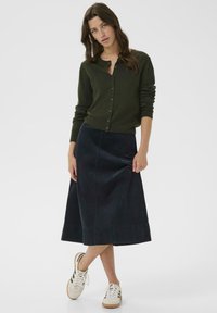 Temně zelený kardigan s knoflíky, kombinovaný s námořnickou áčkovou sukní z měkké tkaniny. Stylizováno s bílými teniskami pro ležérní vzhled.