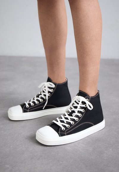 Black high-top canvas sneakers with white rubber soles, featuring white laces and stitching, designed for casual wear.