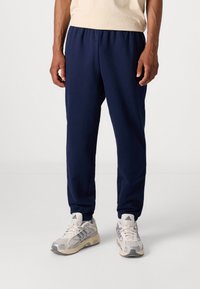 Pantaloni de jogger albaștri, cu talie elastică și glezne strânse, confecționați dintr-un material moale, asociați cu adidași de culoare gri deschis cu accente negre.