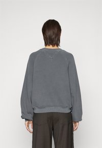 En person med axellångt mörkt hår bär en lös grå sweatshirt och mörka byxor, vänd bort mot en enkel vit bakgrund.