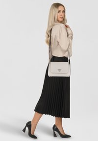 Borsa a tracolla in pelle grigio chiaro con chiusura a patta e logo triangolare, dotata di tracolla staccabile, indossata con una gonna nera plissettata.