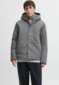 Grå vandtæt jakke med hætte, der har en frontlomme med lynlås og et minimalistisk design. Stoffet har en glat tekstur og en mat overflade.