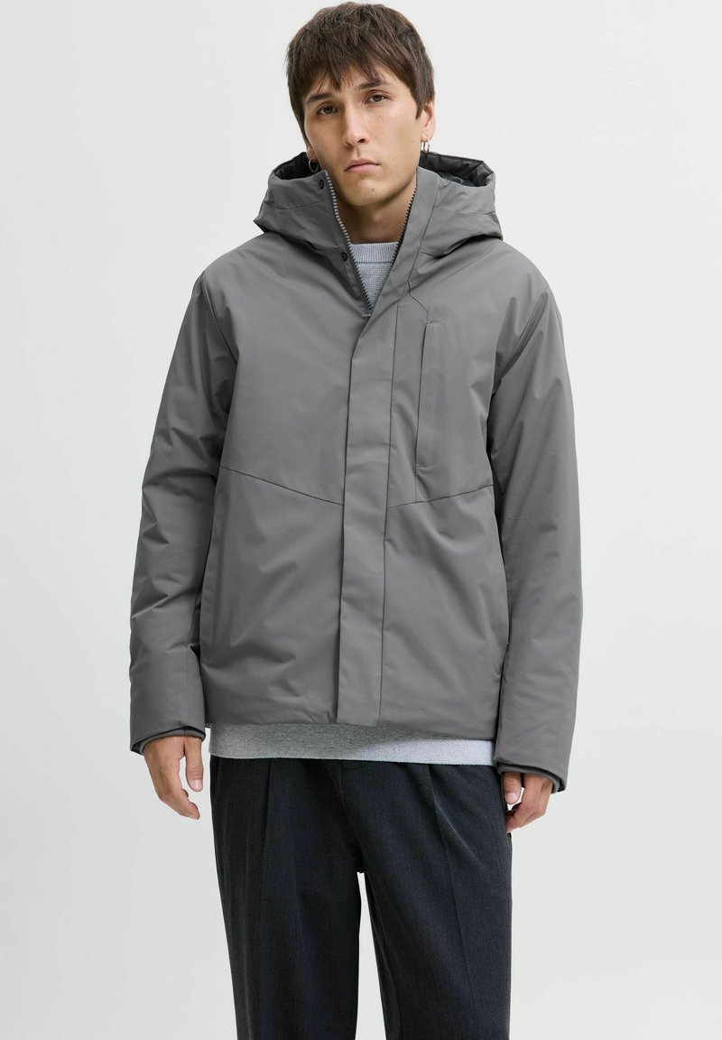 Grå vandtæt jakke med hætte, der har en frontlomme med lynlås og et minimalistisk design. Stoffet har en glat tekstur og en mat overflade.