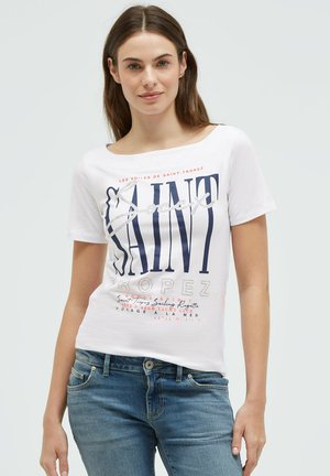 Junge Frau trägt weißes T-Shirt mit Saint-Tropez-Grafik und blaue Jeans, steht vor einfachem hellem Hintergrund und schaut mit leichtem Lächeln nach vorne.