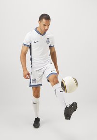Camiseta de fútbol blanca con detalles en azul oscuro, mangas a rayas y logo; combinada con pantalones cortos blancos, calcetas hasta la rodilla y zapatillas negras.