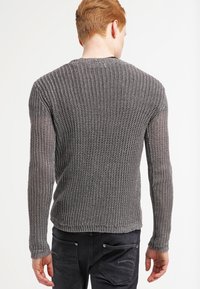 Maglione grigio lavorato a maglia con un design a trama aperta e testurizzata, maniche lunghe e orlo inferiore aderente, indossato con pantaloni scuri.