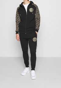 Zwart trainingspak met gouden geometrische patronen op de mouwen en logo. Het jack heeft een vooraanstaande rits; de broek heeft een logo-embleem op de dij.