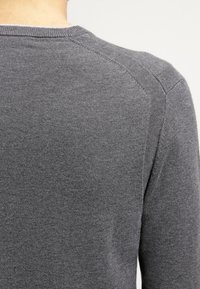 Grå stickad tröja med rundad halsringning, texturerad tyg och raglanärmsdesign. Bakifrån visas en slät, sömlös finish.