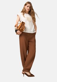 Weißes Kurzarmoberteil mit einem Häkelherz-Detail, kombiniert mit lockeren braunen Hosen und Leopardenprint-High Heels. Trägt eine braune Handtasche.