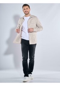 Helles beiges Leinenhemd mit zwei Brusttaschen, über einem weißen T-Shirt unknöpfen, kombiniert mit schwarzen Jeans und weißen Sneakers.
