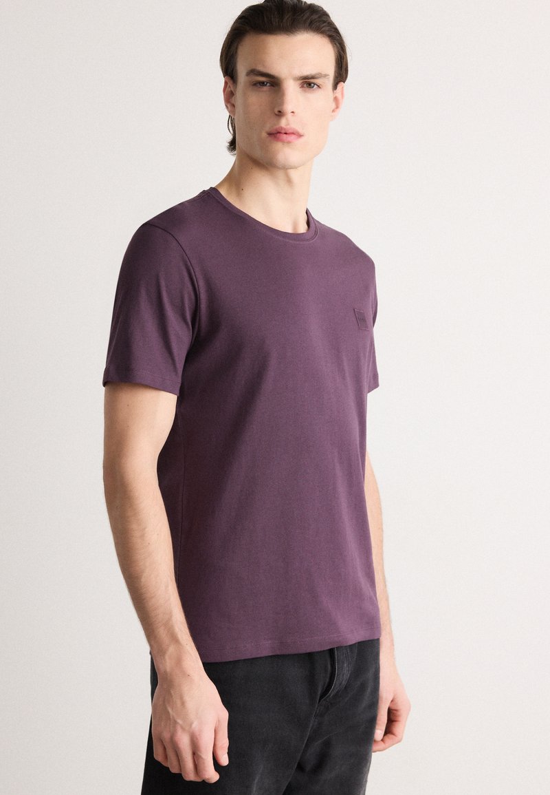 Tricou scurt violet din bumbac moale, cu decolteu rotund și un mic logo pe piept, asortat cu blugi închiși.
