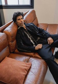 Veste noire texturée avec des accents blancs, chemise noire et pantalon, assis sur un canapé en cuir marron avec un coussin couleur rouille.