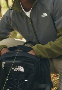 Giacca in fleece grigia e verde oliva con zip, caratterizzata da un logo. Zaino nero con logo e dettagli della cerniera, appoggiato a terra.