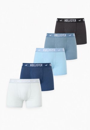 Un mannequin masculin se tient devant un fond clair uni, vêtu d’un caleçon boxer noir Hollister.