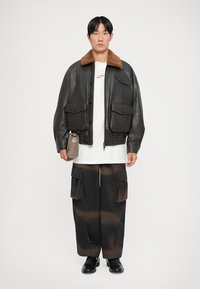Black leather bomber jacket with a brown fur collar, featuring two chest pockets, paired with loose dark cargo pants and black shoes.