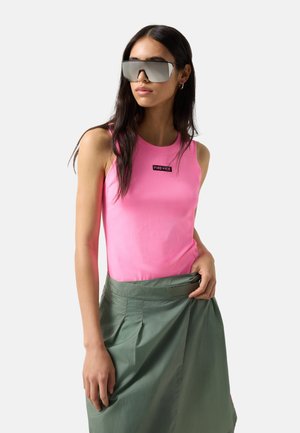 Kvinde iført store reflekterende solbriller, en lyserød ærmeløs top med "FIRE + ICE" mærkat og et grønt skørt, poserende foran en ensfarvet baggrund.