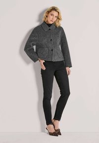 Vrouw in een grijs knoopjesshirt, zwarte skinny jeans en hoge hakken met panterprint, staand tegen een effen lichte muur.