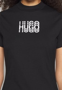 Czarna bawełniana koszulka z krótkim rękawem, z wyraźnym białym logo "HUGO" w stylizowanym, falistym tekście. Wysoki dekolt.