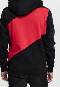 Osoba nosící černou a červenou mikinu s kapucí s diagonálním červeným panelem přes záda, doplněnou o černé džíny.