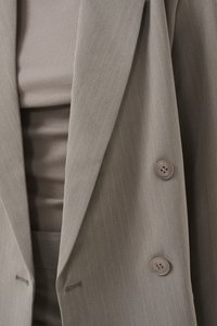 Sacou tailored de culoare gri deschis, cu dungi verticale, dotat cu două butoane și rever în formă de v. Sub acesta, se vede un top bej cu dungi.