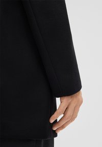 Cappotto nero con texture e design slim. Le maniche sono leggermente sovradimensionate, con una mano visibile vicino all'orlo. Nessuna ferramenta o dettagli visibili.
