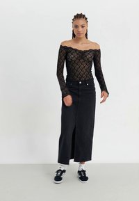 Junge Frau mit geflochtenem Haar, die ein schwarzes Off-Shoulder-Spitzenoberteil, einen langen schwarzen Rock mit Schlitz vorne, weiße Socken und schwarze Turnschuhe vor einem weißen Hintergrund trägt.