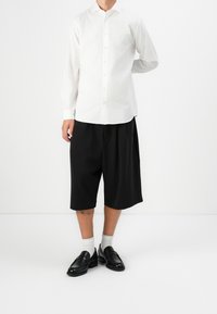 Camicia bianca a maniche lunghe con chiusura a bottoni abbinata a pantaloni corti neri larghi. Mocassini neri e calzini grigi chiari completano l'outfit.