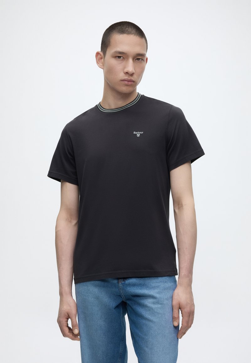 Νεαρός άνδρας φοράει μαύρο t-shirt Barbour με ριγωτή λεπτομέρεια στο γιακά και ανοιχτό μπλε τζιν, στέκεται μπροστά σε απλό λευκό φόντο.