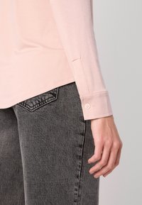 Lys rosa skjorte med lang erme og knappede mansjetter, sammen med mørk grå denim jeans som har et karakteristisk lomme design.