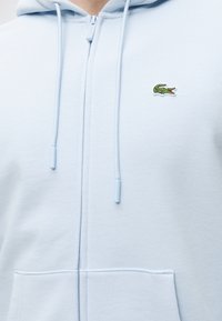 Sweat à capuche zippé bleu clair avec cordons et petit logo crocodile vert sur la poitrine gauche.