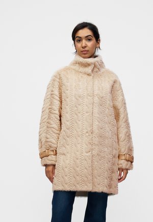 Mujer con un abrigo de piel texturizado beige con cuello alto y detalles de hebillas en las mangas, combinado con pantalones azul oscuro.