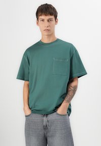 Green cotton t-shirt with a round neck, short sleeves, and a chest pocket. The fabric has a smooth texture and is paired with gray jeans.