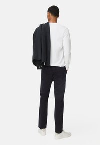 Witte lange mouwen shirt, donkere jas over de schouder gedrapeerd, donkere broek en lichte sneakers. Model staat met de rug naar de camera tegen een neutrale achtergrond.