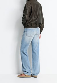 Personne portant une veste en cuir marron, un jean large bleu clair et des sandales beiges à clous, se tenant contre un mur blanc.