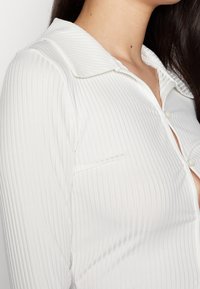 Camicia bianca con colletto, a righe verticali, dotata di bottoni e taschino frontale. Il tessuto appare liscio e leggermente trasparente.