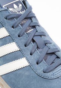 Blue suede sneaker with white stripes, round laces, and a cushioned tongue. Features textured fabric and a logo tag on the side.