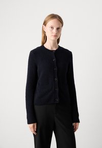 Frau mit glattem blonden Haar trägt Creolen, eine dunkelblaue, geknöpfte, strukturierte Strickjacke und schwarze Hosen vor einem schlichten weißen Hintergrund.