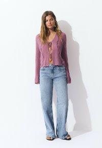 Purple cropped knit sweater with V-neck and tie detail, paired with light blue high-waisted flared jeans. Textured fabric, casual fit.
