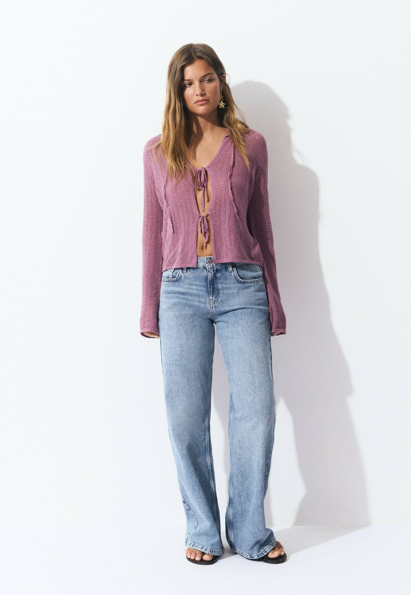 Purple cropped knit sweater with V-neck and tie detail, paired with light blue high-waisted flared jeans. Textured fabric, casual fit.