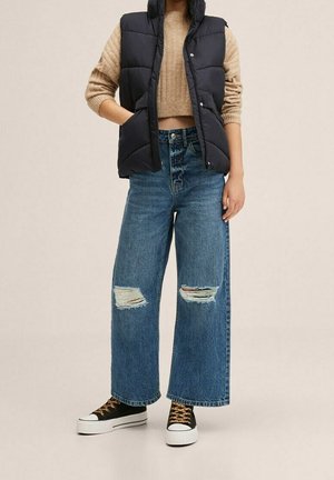 Person iført en tanfarvet cropped sweater, sort dunvest, revnede blå wide-leg jeans og sorte høje sneakers med leopardprint-snørebånd.