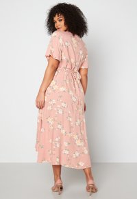 Kopertowa sukienka midi w delikatnym różowym kolorze, z krótkimi rękawami, wiązanym w pasie oraz falbaniastym dołem, ozdobiona białymi i żółtymi wzorami kwiatowymi.