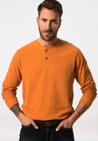 Oranje lange mouwen shirt met een ribbelstructuur, voorzien van een ronde halslijn en een tweeknoopsluiting. Gecombineerd met donkere jeans en een bruine riem.
