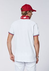 Weißes Polohemd mit rotem Kragen, versehen mit Text und Logo. Rote Kappe auf dem Kopf des Models. Kurze Ärmel mit marineblauen und roten Akzenten.