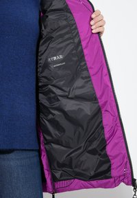 Ruka držící otevřenou fialovou bundu s černou podšívkou, na které je logo "HYRAR" a označení odpudivosti vody, nošenou přes modrý svetr a džíny.