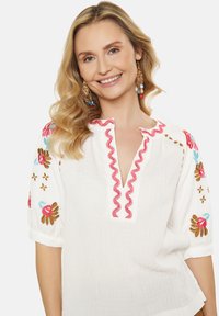 Bluza albă texturată cu broderie florală în roșu, albastru și maro, având un decolteu în V, cu margine de rickrack roz și mâneci scurte tip puff.