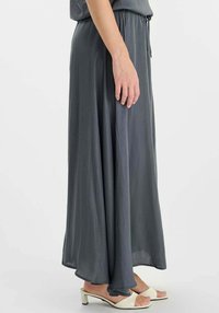 Lange, A-lijn rok van zachte, glinsterende grijze stof. Heeft een elastische tailleband en is halflang, gedragen met witte open schoenen.