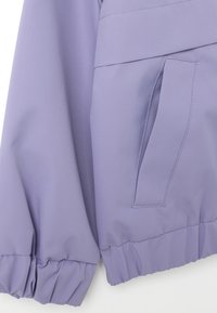 Giacca leggera lilla con vita aderente, polsini elastici e una tasca laterale con una leggera lucentezza. Tessuto morbido senza motivi visibili.