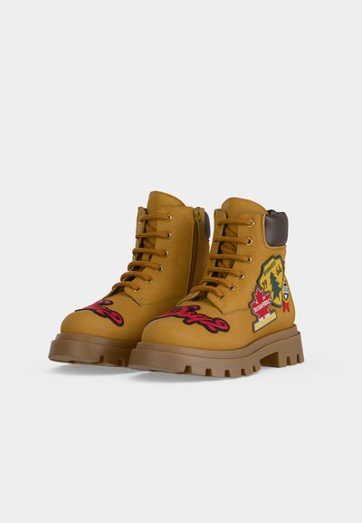 Paire de bottes lacées couleur fauve avec semelles épaisses, fermeture éclair latérale, col matelassé et écussons brodés colorés sur la pointe et les côtés extérieurs.